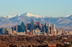 view of the Denver, Colorado skyline at golden hour, with snow-capped Rocky Mountains in the background and a light layer of snow on rooftops and trees in the foreground
